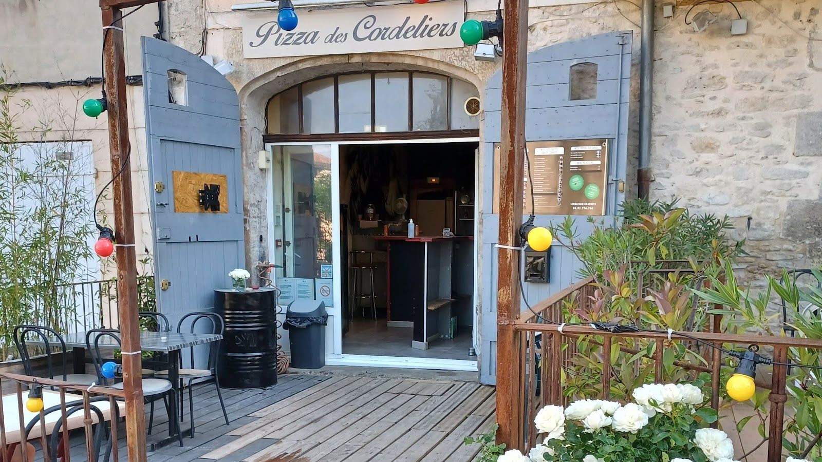 La devanture de la pizzeria : volets bleu-gris ouverts, guirlande d'ampoules colorées, terrasse en bois